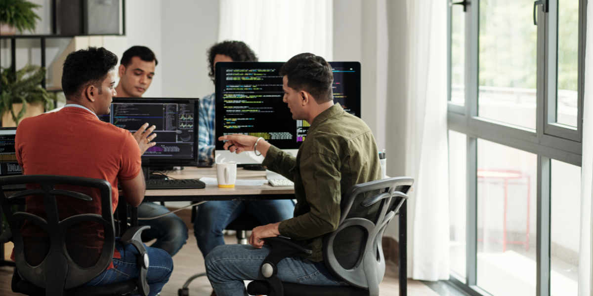 Four IT technicians working together at desktop computers with programming code on screens in modern office with natural lighting